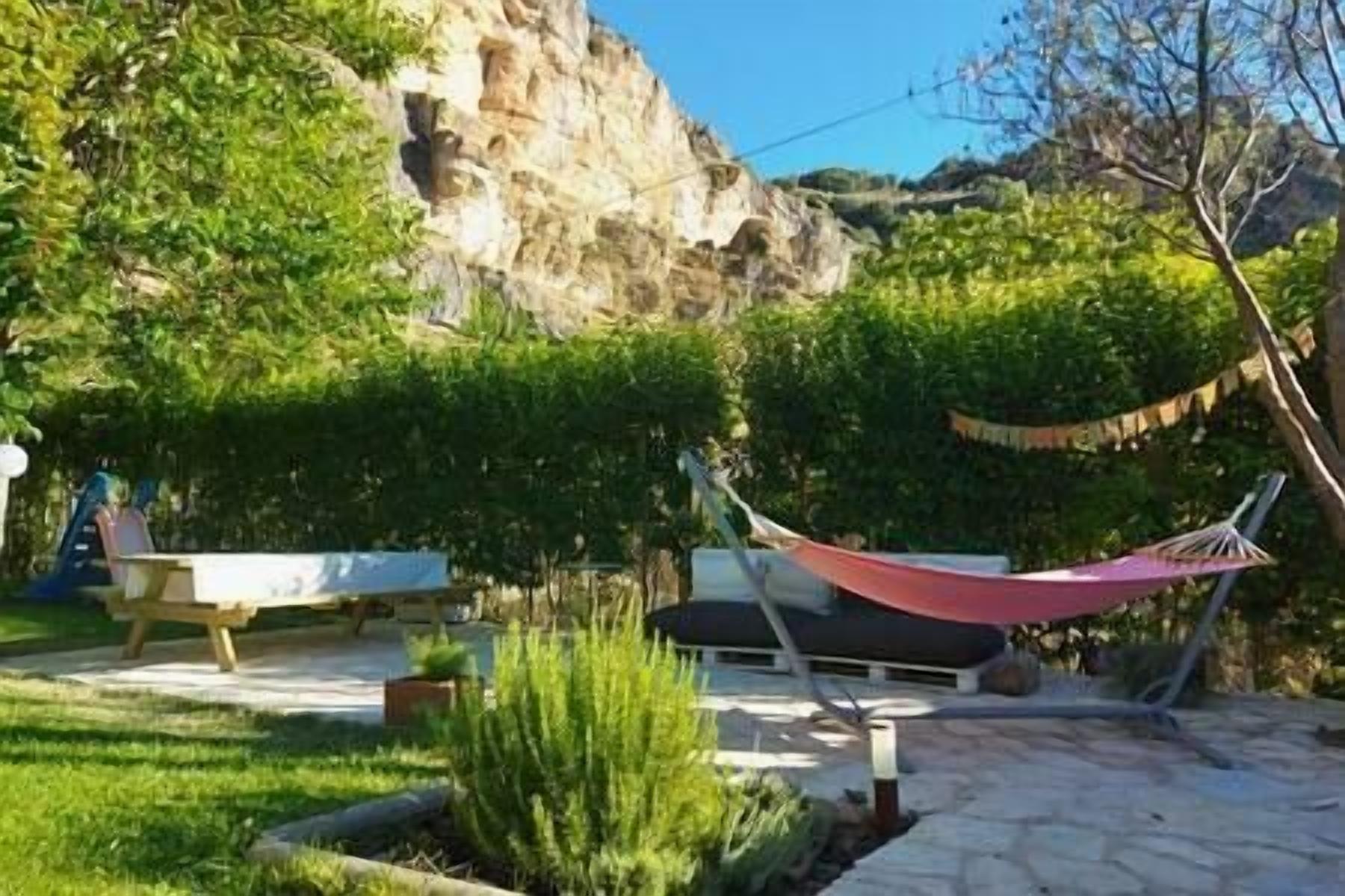 A patio area with a hammock, a bench, and a small table. There is a rocky cliff in the background and a tree to the right.