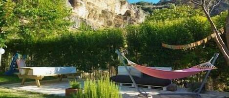 A patio area with a hammock, a bench, and a small table. There is a rocky cliff in the background and a tree to the right.