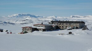 A snow-covered mountain landscape with a building featuring a stone facade and a red roof. - Rødungstøl Høyfjellshotell (Al)