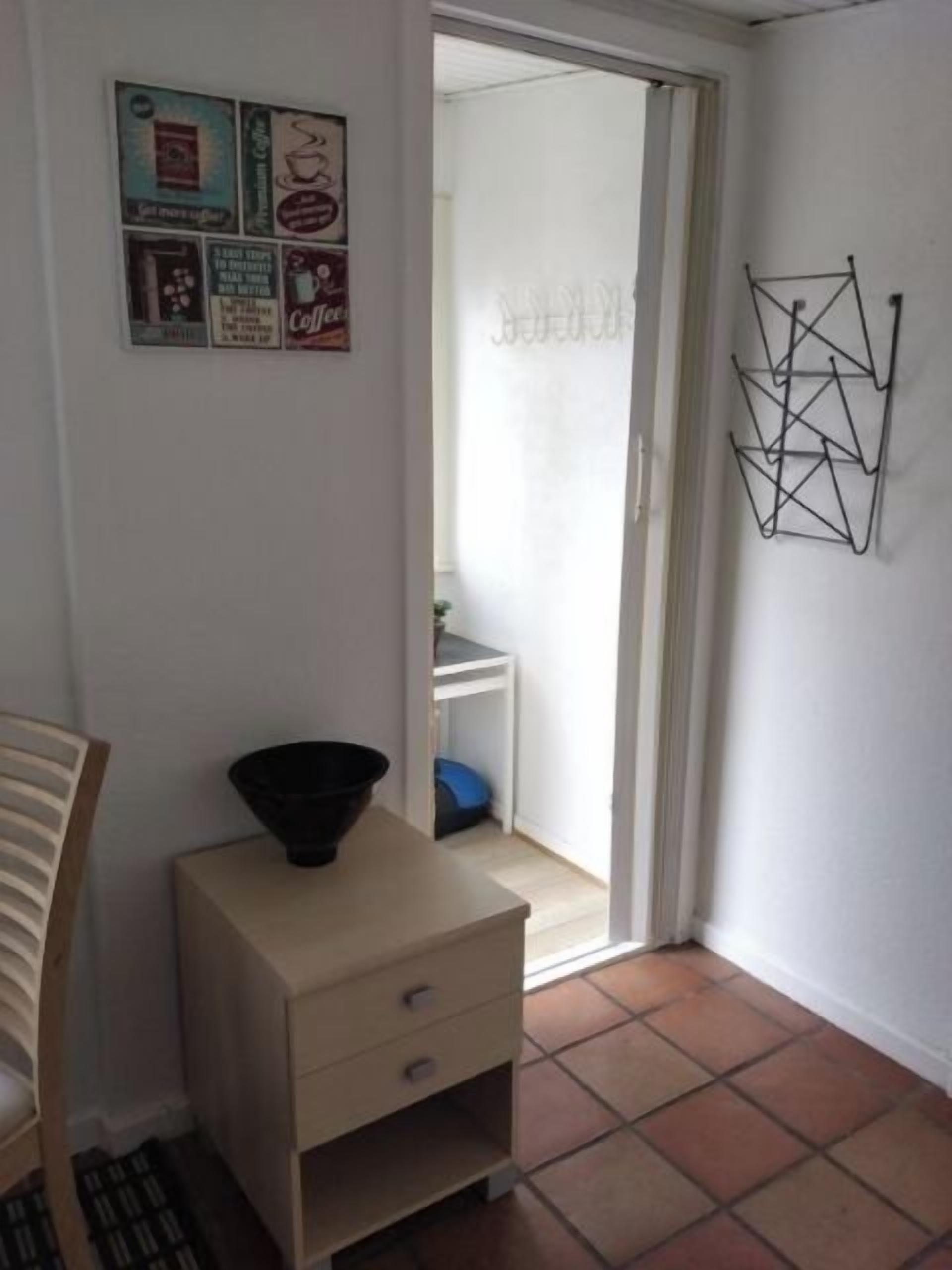Una habitación pequeña con una pared blanca, una puerta que da a un balcón, una mesa pequeña con un tazón negro y una silla.
