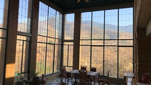 Amplia habitación con ventanas grandes que ofrecen vistas a las montañas. Cuenta con un ventilador de techo, una mesa de comedor con sillas y varias plantas en macetas.
