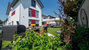 A modern two-story house with a white exterior and red accents, a visible garage, and a well-maintained garden with outdoor seating. - Design apartment for 3-5 people with south-facing loggia and garden view. Near the lake! (Bensheim)