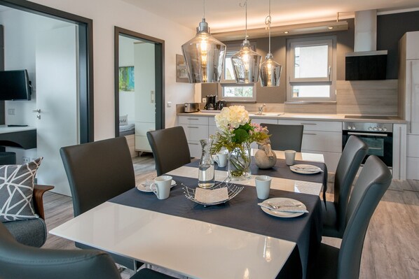 A modern dining area with a table set for four, surrounded by chairs. There are hanging pendant lights above the table, and a vase with flowers on the table.
