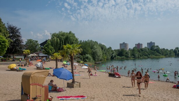 A beach with people swimming and sunbathing, surrounded by trees and buildings. - Design apartment for 3-5 people with south-facing loggia and garden view. Near the lake! (Bensheim)