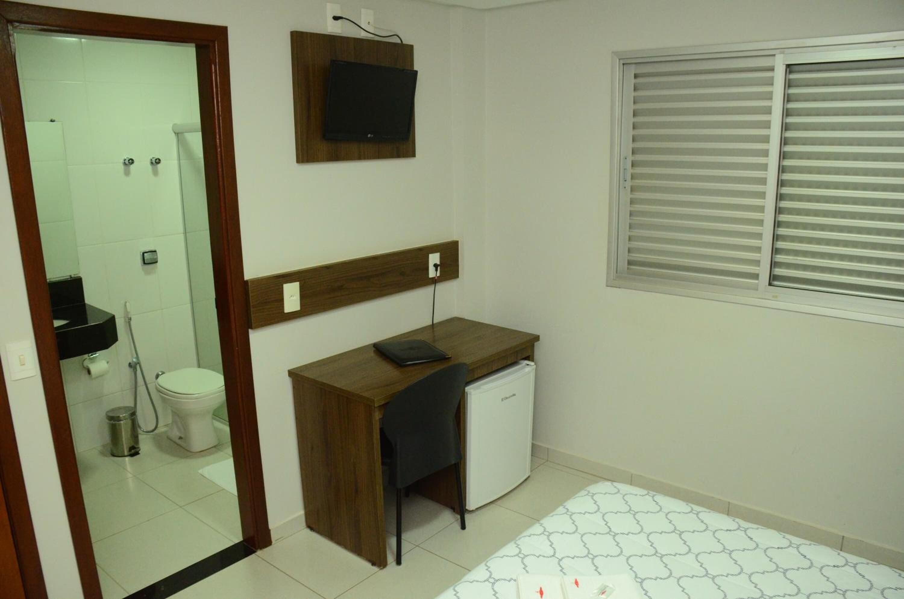 Una habitación compacta con escritorio de madera, un refrigerador pequeño, inodoro y ventana con persianas.