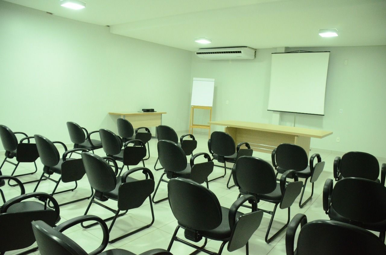 Un salón de conferencias con filas de sillas negras, un atril, una pizarra y un equipo de aire acondicionado en la pared.