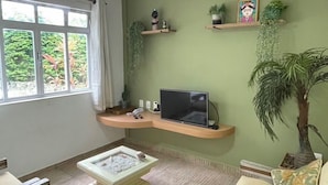 Ein Wohnzimmer mit einer Couch, einem Couchtisch, einem Fernseher, einem Deckenventilator und einem Fenster mit Blick ins Grüne.