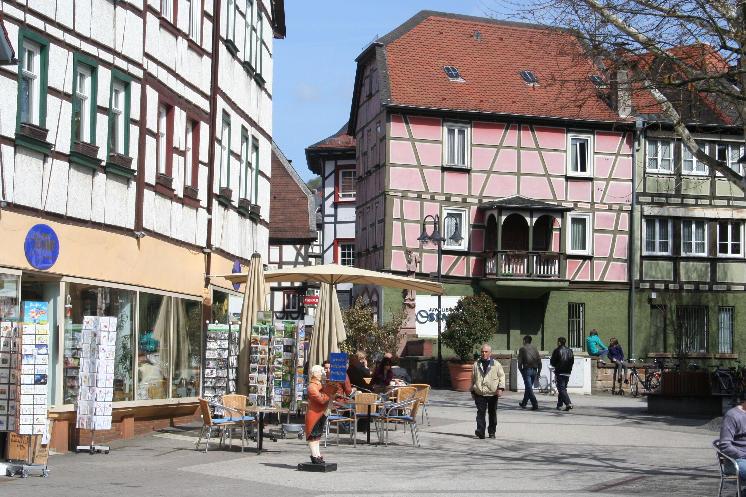 A traditional European street with half-timbered buildings, outdoor seating, and a statue.