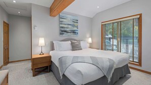 Une chambre avec un grand lit, deux tables de chevet avec des lampes, une poutre en bois au plafond, une fenêtre avec des stores et un tableau au mur.