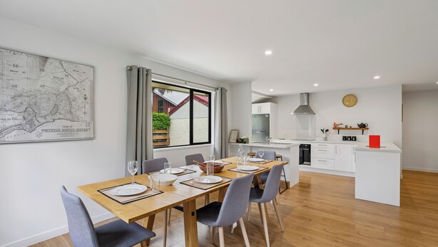 A modern kitchen with white cabinets, a central island, and a dining area with a wooden table and grey chairs. - Rees on Reid - Arrowtown Holiday Home (Arrowtown)