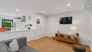 A modern living room with a grey sofa, wooden flooring, a TV mounted on the wall, a wooden cabinet, and a window with a view of greenery. - Rees on Reid - Arrowtown Holiday Home (Arrowtown)