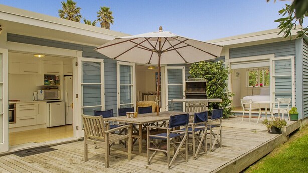 Une terrasse en bois avec une table et des chaises pour les repas, un barbecue et un petit coin cuisine visible à travers une porte ouverte.