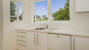Une cuisine avec des meubles blancs, un évier en acier inoxydable et deux fenêtres donnant sur un paysage verdoyant.