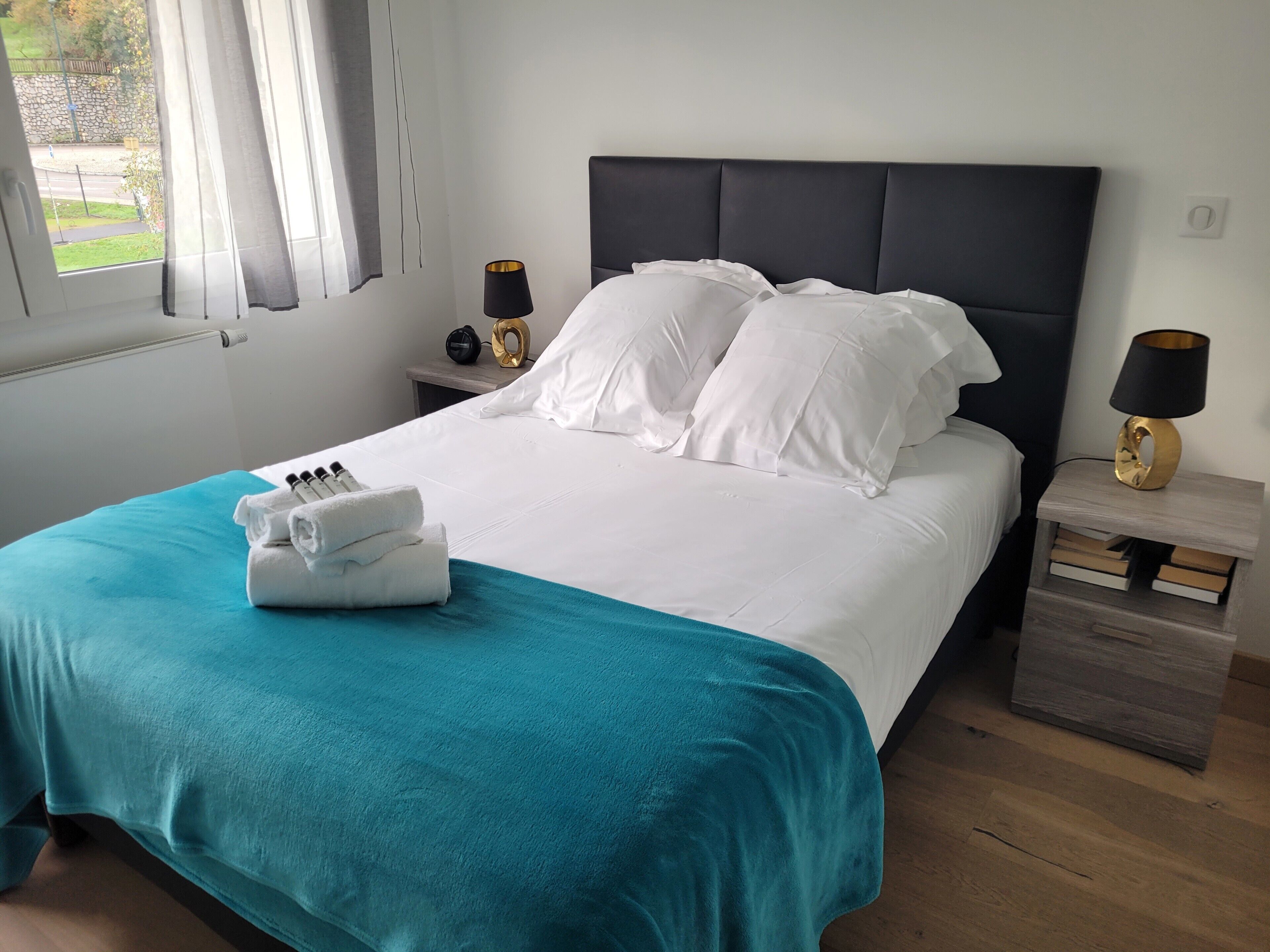 A neatly made bed with white linens and a teal throw blanket, a bedside table with a lamp, and a view of the outdoors through a window.