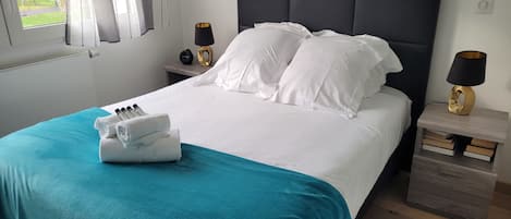 A neatly made bed with white linens and a teal throw blanket, a bedside table with a lamp, and a view of the outdoors through a window.