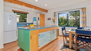 A modern kitchen with a central island, wooden cabinets, and a view of the outdoors. - Taste Of Paradise with WiFi!- Mangawhai Heads Home (Mangawhai Heads)