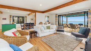 A spacious living area with a large window offering a view of the sea, a dining area with a green table, and a cozy seating arrangement with wicker furniture and a gray rug. - Taste Of Paradise with WiFi!- Mangawhai Heads Home (Mangawhai Heads)