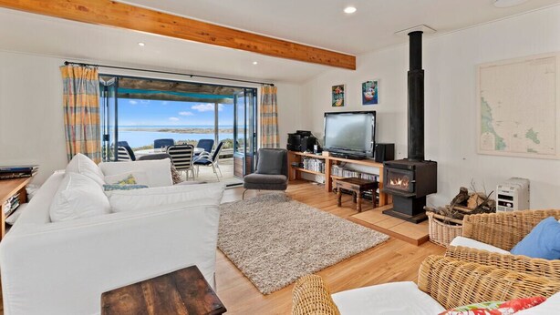 A cozy living room with a white sofa, a television, a fireplace, and a view of the sea through large windows. - Taste Of Paradise with WiFi!- Mangawhai Heads Home (Mangawhai Heads)