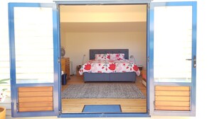 A bedroom with a bed, a bedside table, a lamp, and a wooden floor. - Taste Of Paradise with WiFi!- Mangawhai Heads Home (Mangawhai Heads)