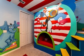 Un cuarto infantil con un mural de un personaje de gato, una puerta y una zona de juegos circular con un tobogán.