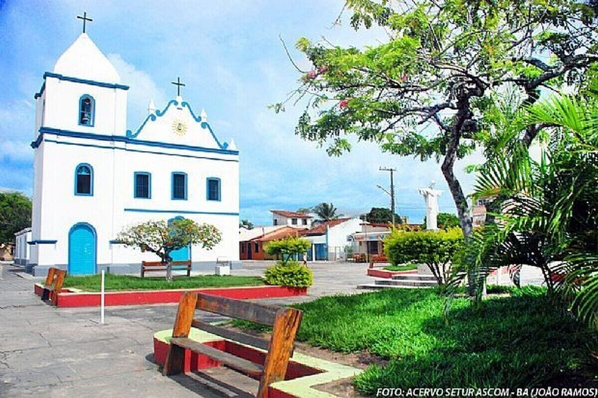 Una chiesa bianca con una croce blu sulla sommità, immersa nel verde e circondata da panchine.