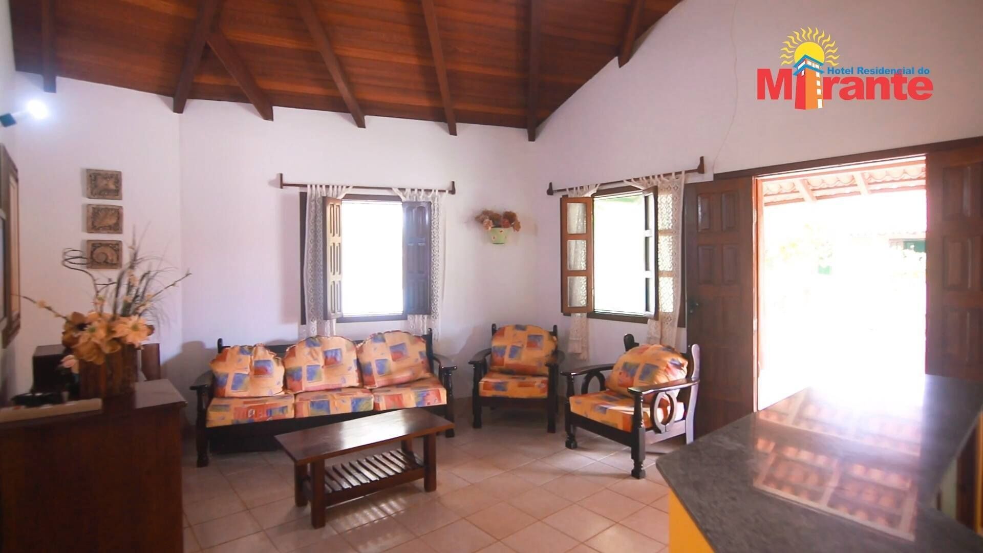Una sala de estar con un sofá, una mesa de centro de madera y dos sillones. Hay ventanas con cortinas, una puerta que da al exterior y un espejo montado en la pared.