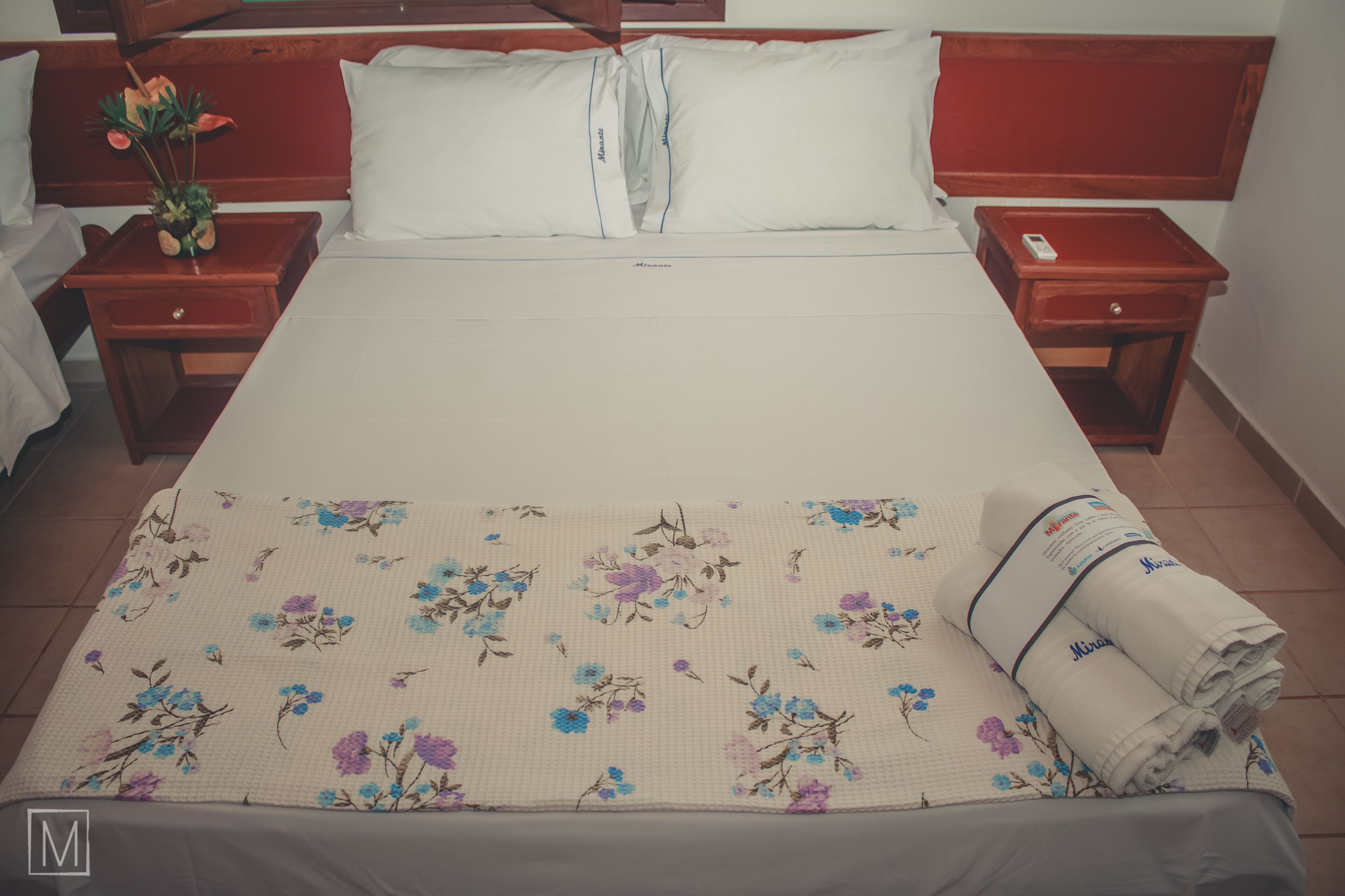 Een keurig opgemaakt bed met een bloemenbedekking, twee nachtkastjes met lampen, en een nachtkastje met een afstandsbediening.