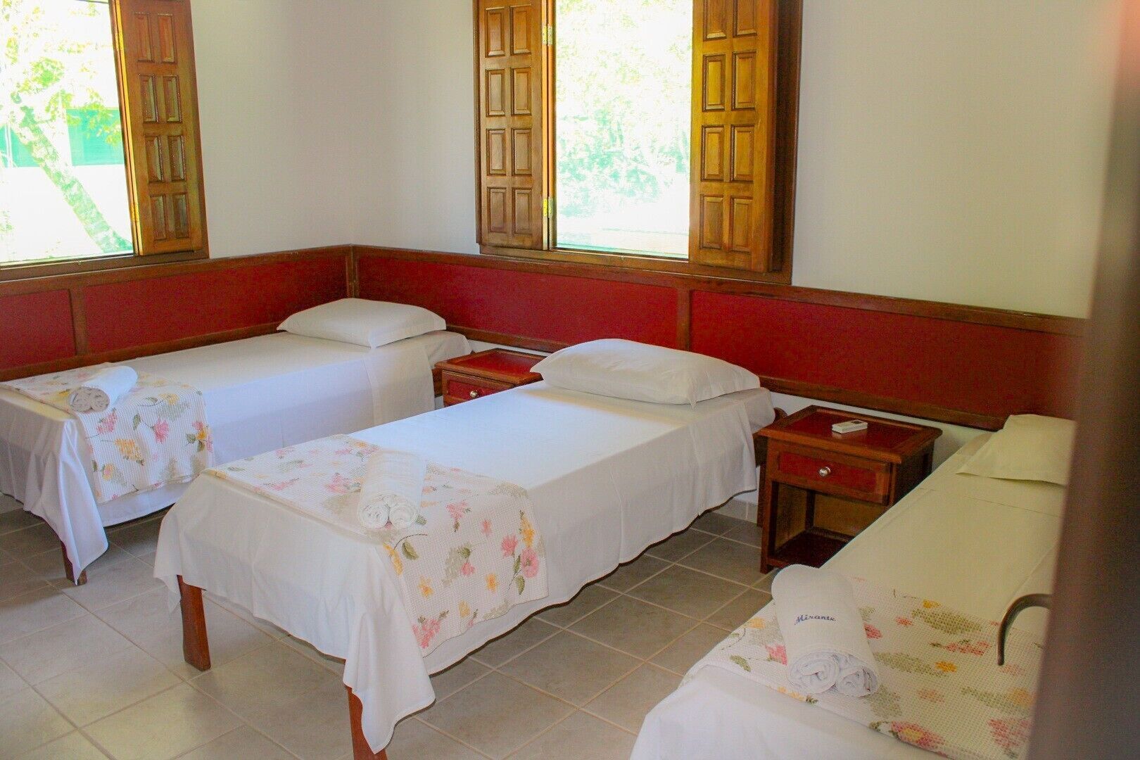 Habitación de hotel con dos camas individuales, cabecera de madera, mesita de noche pequeña con lámpara y ventana con vista a zonas verdes.