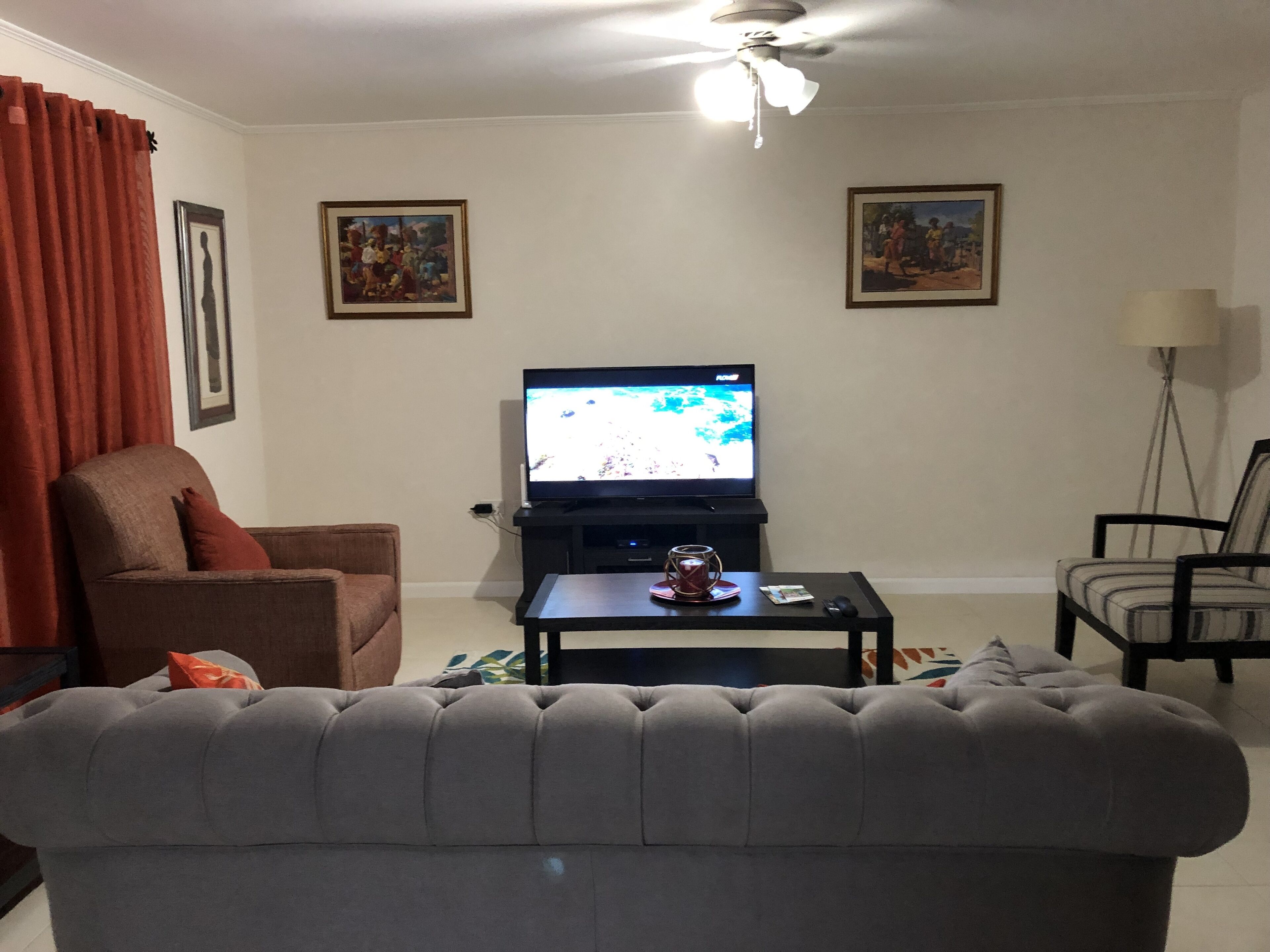 A living room with a grey sofa, a brown armchair, a black coffee table, and a television on a stand. There are two framed pictures on the wall and a floor lamp to the right.