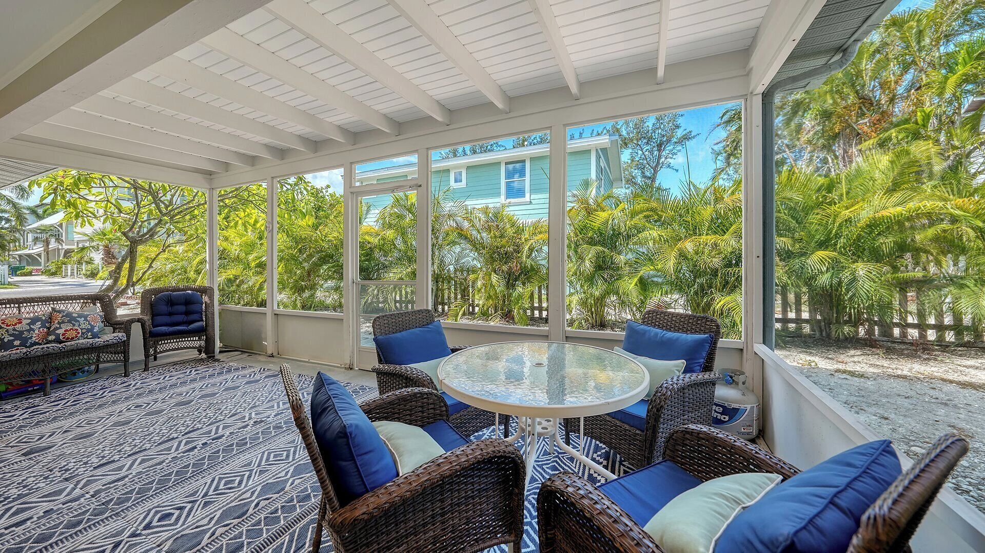 A sunlit covered patio with wicker furniture, a glass table, and a view of a blue house and tropical vegetation.