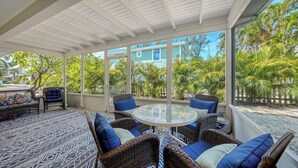 Een zonnig overdekt terras met rieten meubels, een glazen tafel en uitzicht op een blauw huis en tropische begroeiing.