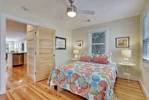 A bedroom with a wooden floor, a bed with a colorful floral bedspread, two bedside lamps, and a ceiling fan. - Classic Folly Cottage (Folly Beach)