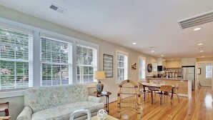 A spacious living room with a sofa, dining table, and chairs, large windows with blinds, and a kitchen area in the background. - Classic Folly Cottage (Folly Beach)