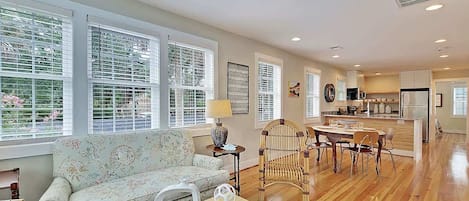 A spacious living room with a sofa, dining table, and chairs, large windows with blinds, and a kitchen area in the background.