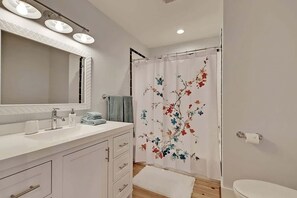 A bathroom with a white vanity, a large mirror, a shower curtain with a floral design, and a toilet. - Classic Folly Cottage (Folly Beach)