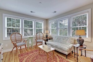 Ein großzügiges Wohnzimmer mit Sofa, zwei Sesseln, einem Couchtisch und einem Teppich. Große Fenster mit Jalousien sorgen für viel Tageslicht.