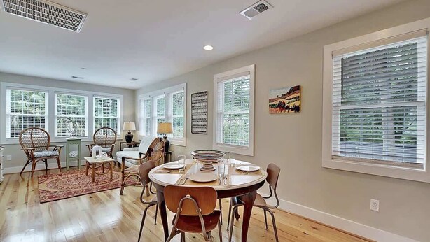 A dining area with a round table set for four, surrounded by wooden chairs. There are large windows with blinds, a decorative rug, and a wall-mounted artwork. - Classic Folly Cottage (Folly Beach)