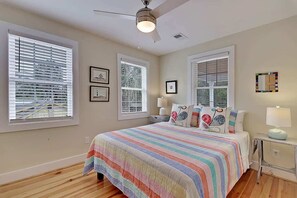 Une chambre avec un grand lit, de nombreuses fenêtres, un ventilateur de plafond et des tableaux encadrés accrochés au mur.