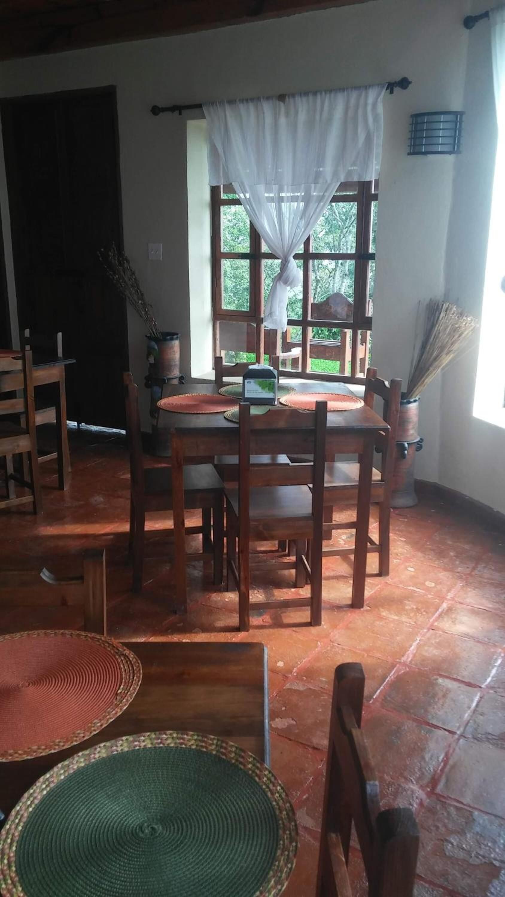 A dining area with a wooden table and chairs, a window with a view of greenery, and a rustic tiled floor.