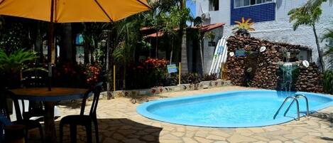 A swimming pool with a stone patio, a yellow umbrella, and a small waterfall feature.