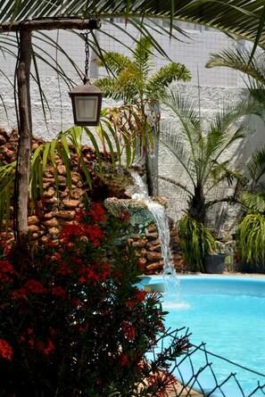 Piscina com cachoeira, rodeada por plantas tropicais e uma parede de pedra.