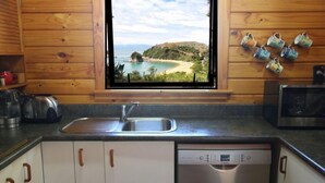 A kitchen with wooden cabinets, a dishwasher, and a window with a view of a beach and mountains. - Heart of Kaiteriteri: Absolute Privacy nestled between both beaches  (Kaiteriteri)