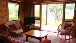 A cozy room with a sofa, two armchairs, a coffee table, a TV, and large windows offering a view of trees. - Heart of Kaiteriteri: Absolute Privacy nestled between both beaches  (Kaiteriteri)