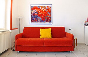En röd soffa med en gul kudde i ett rum med ett vitt element, en inramad abstrakt målning på väggen och ett litet bord med en lampa.