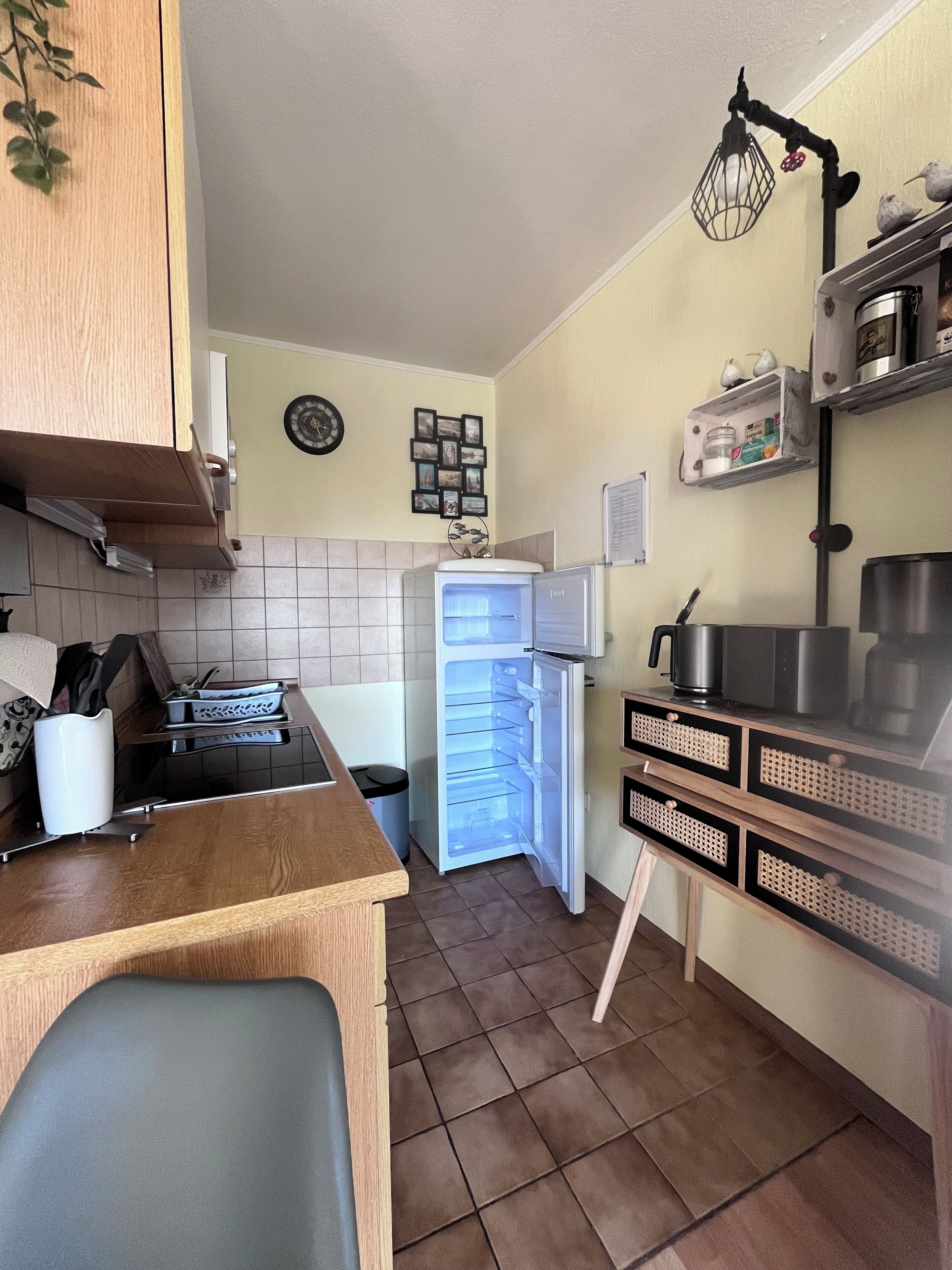 Eine kompakte Küche mit integriertem Kühlschrank, Herd und Spüle. Eine kleine Ablagefläche zeigt dekorative Gegenstände, an der Wand hängt eine Uhr.