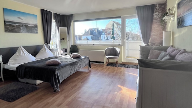 A modern living room with a bed, sofa, and a chair. There is a window with curtains, a chandelier, and artwork on the walls. - Cozy apartment, Cuxhaven-Döse (Cuxhaven)