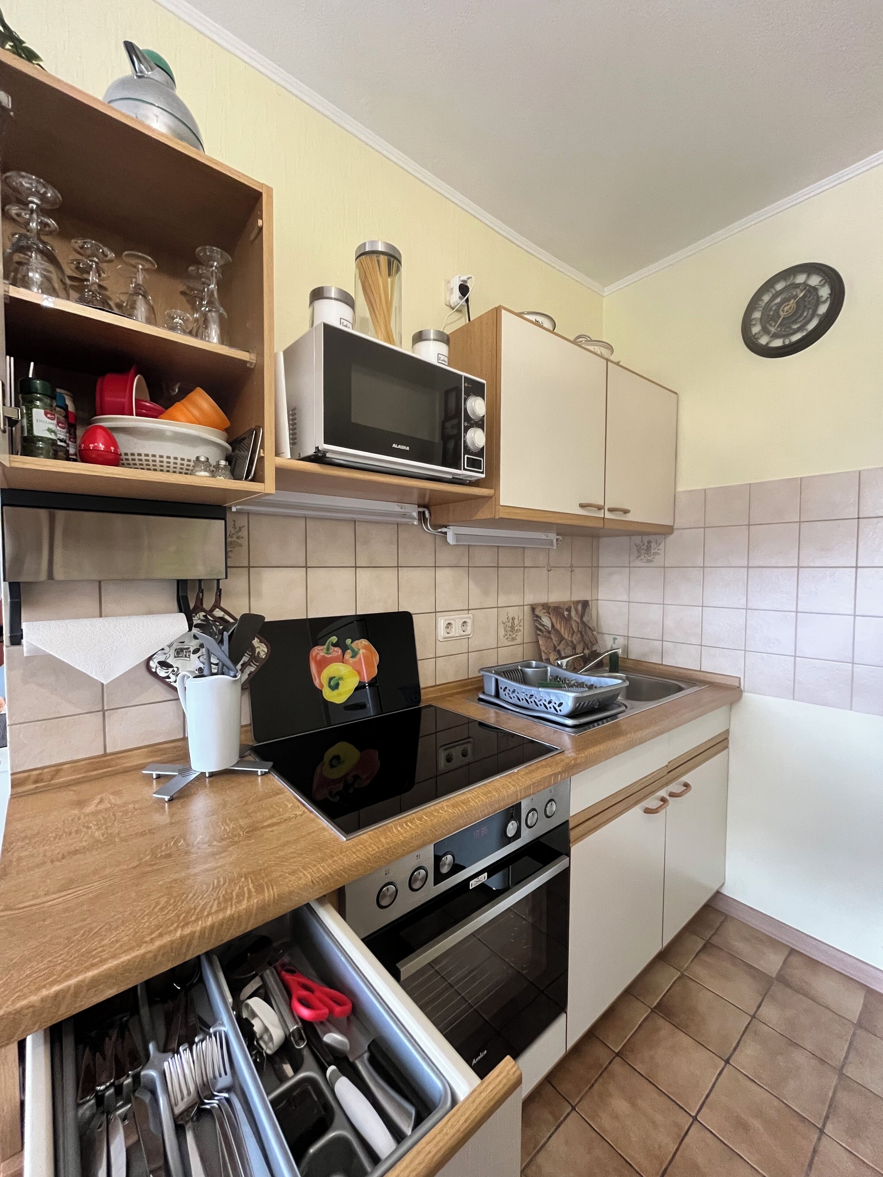 A kitchen with a microwave, stove, oven, and a built-in sink. There is a shelf with glassware and a clock on the wall.