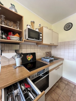 A kitchen with a microwave, stove, oven, and a built-in sink. There is a shelf with glassware and a clock on the wall. - Cozy apartment, Cuxhaven-Döse (Cuxhaven)