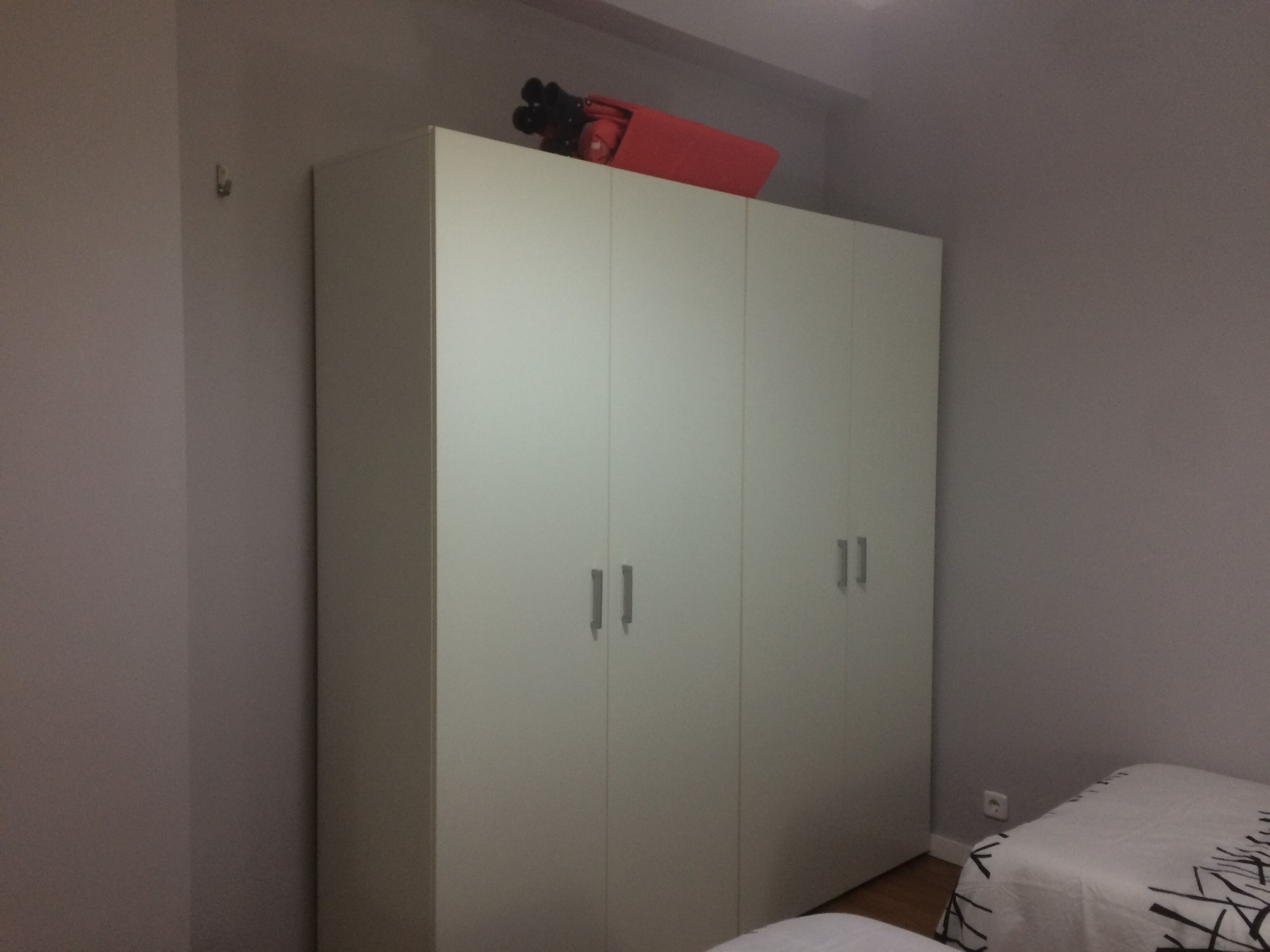 Une chambre avec une grande armoire, un lit recouvert d’une couvre-lit à motifs et un objet rouge posé sur le dessus de l’armoire.