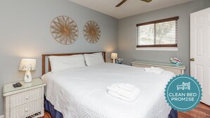 A clean, well-lit bedroom with a bed, two wall decorations, a ceiling fan, a window with blinds, and a bedside table with a lamp. - Waterfront Escape | Pet-Friendly & Beach Nearby (Corpus Christi)